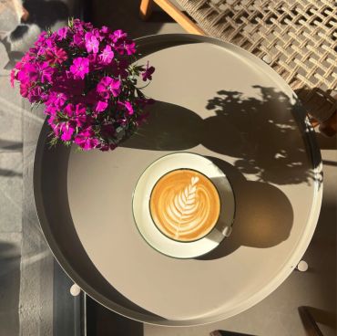 A cup of cappuccino with latte art next to vibrant purple flowers on a round table.