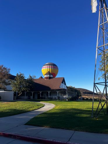 A colorful hot air balloon appears behind a house under a clear blue sky.