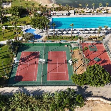 Aerial view of a sports and leisure area with tennis courts, a basketball court, and swimming pools near the sea.