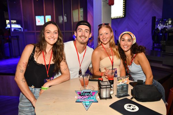 Four friends smiling at a nightlife event with drinks and red lanyards.