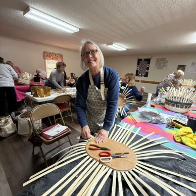 Woman weaving basket strips in a craft workshop with others in the background.