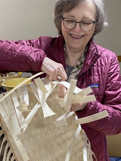 A smiling woman weaving a basket indoors.