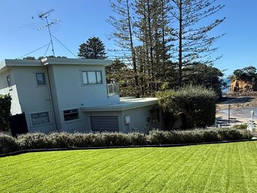 A house with a green lawn overlooking the ocean and rocky shore under a clear blue sky.