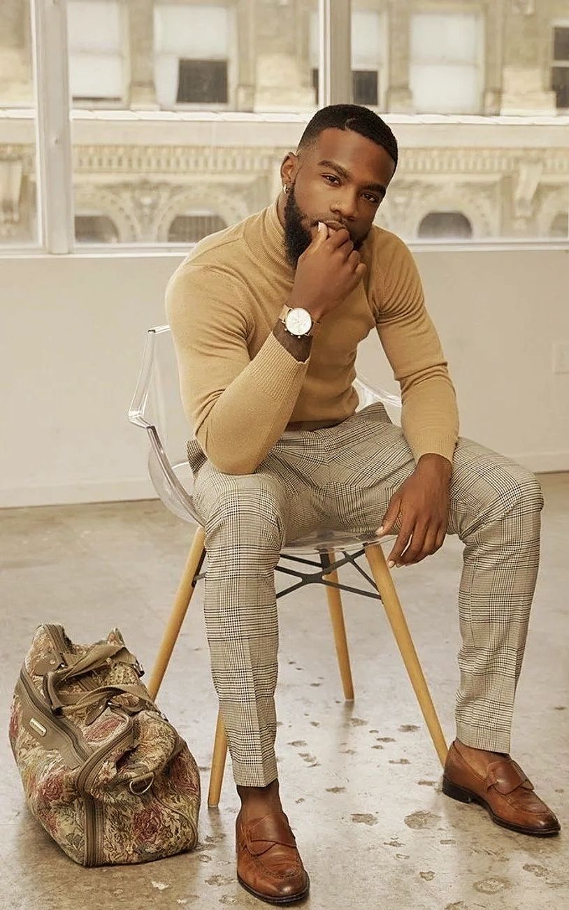 Stylish man seated on a clear chair, wearing a beige turtleneck and plaid pants.