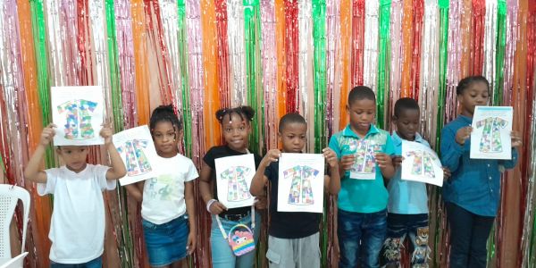 Seven children holding colorful drawings of shirts, standing in front of a festive backdrop.