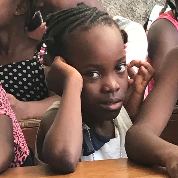 A young girl with braided hair sits at a table, resting her head on her hands.