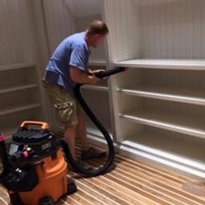 Man vacuuming shelves with a shop vacuum in a room with wooden floors.