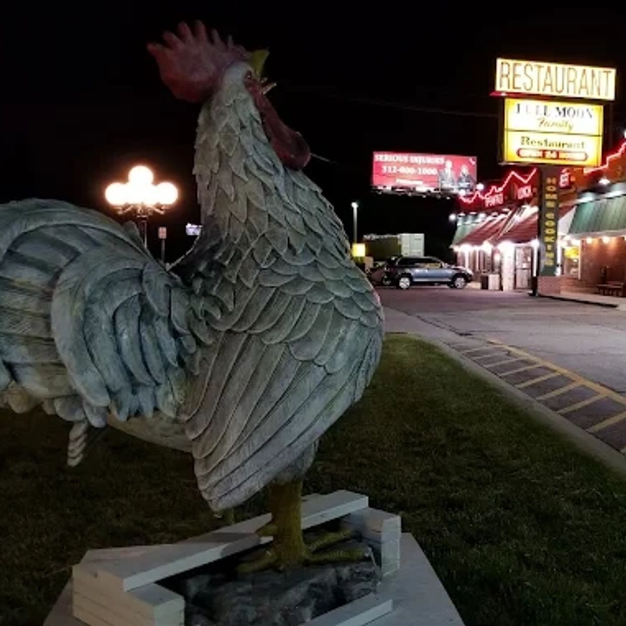 Large metal rooster statue outside a brightly lit restaurant at night.