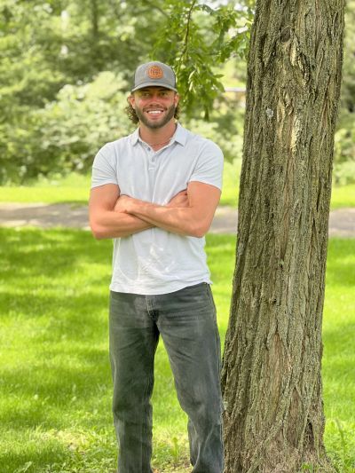 Man in a white polo and cap standing by a tree in a park.