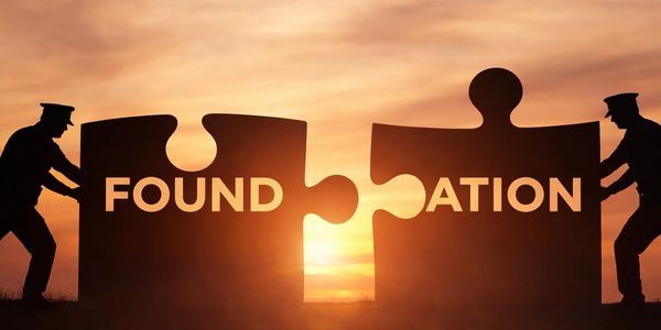 Two silhouetted figures push puzzle pieces forming the word 'FOUNDATION' at sunset.