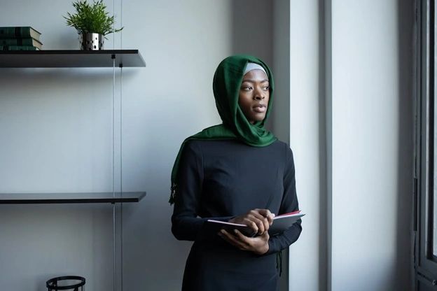 A black woman in a hijab holding a book and looking out the window. A black woman in a hijab holding a book and looking out the window.