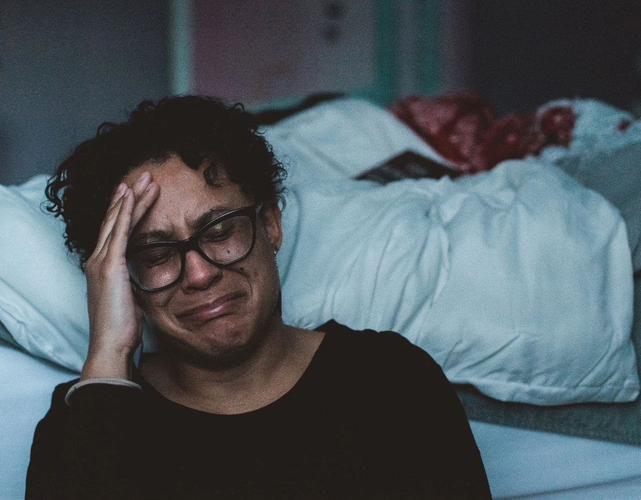 A woman in glasses, leaning up against her bed, crying. A woman in glasses, leaning up against her bed, crying.