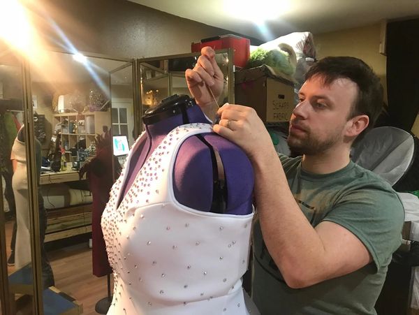 A man adjusts a studded white dress on a mannequin in a workshop.