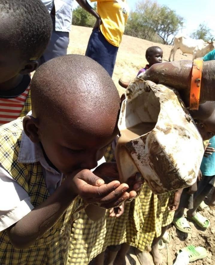 A child drinks water from a container held by an adult in a rural setting.