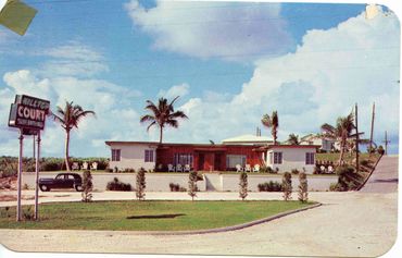 Vintage motel with palm trees, outdoor chairs, and a classic car under a blue sky.