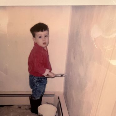 Young child painting a wall indoors, standing on a covered floor surface.