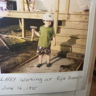 A young boy named Larry holding a hammer, wearing a hard hat and standing in front of a wooden structure.