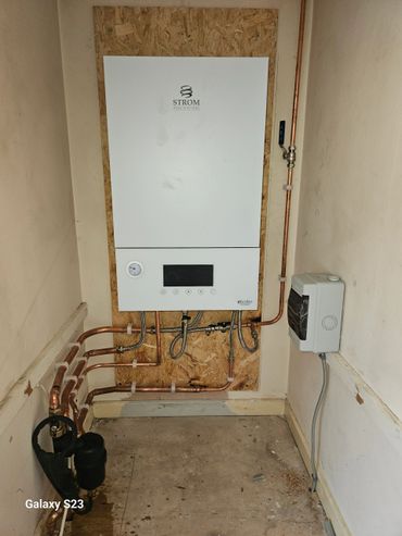 Modern white Strom eBoiler with copper piping installed on wooden board in utility room.