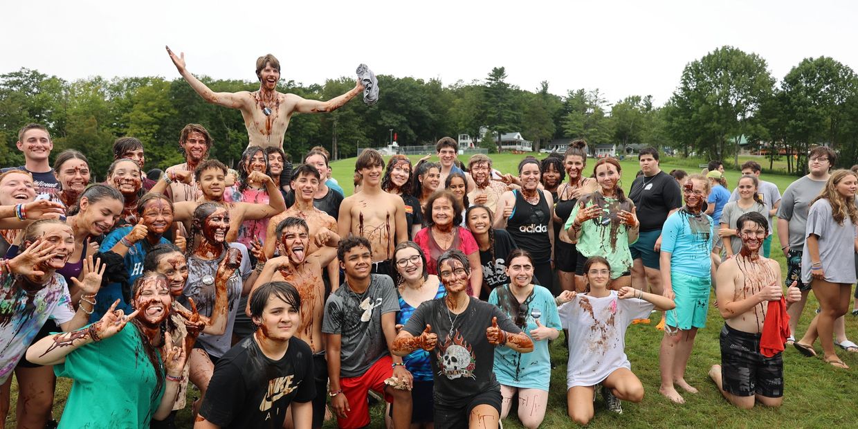 A large group of smiling teenagers covered in mud gathered outdoors on grass.