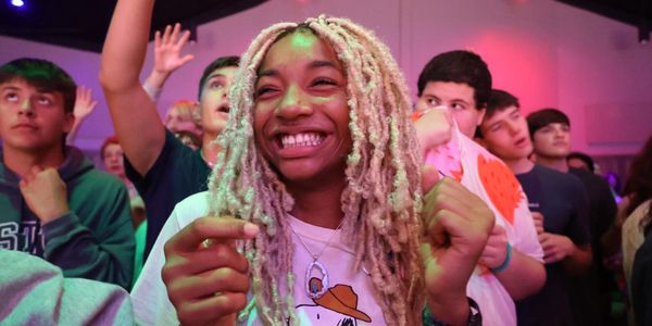 A joyful woman with blonde dreadlocks smiles at a lively event surrounded by an excited crowd.