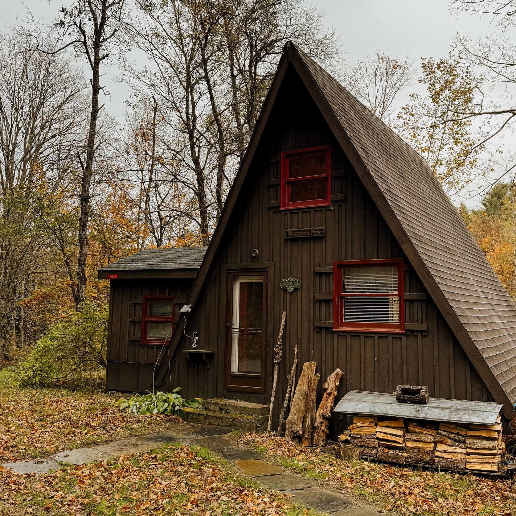 A rustic A-frame cabin surrounded by autumn foliage with stacked firewood outside.