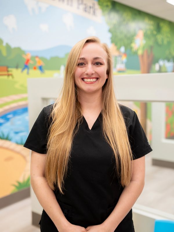 Smiling woman with long blonde hair wearing black scrubs in a colorful indoor setting.