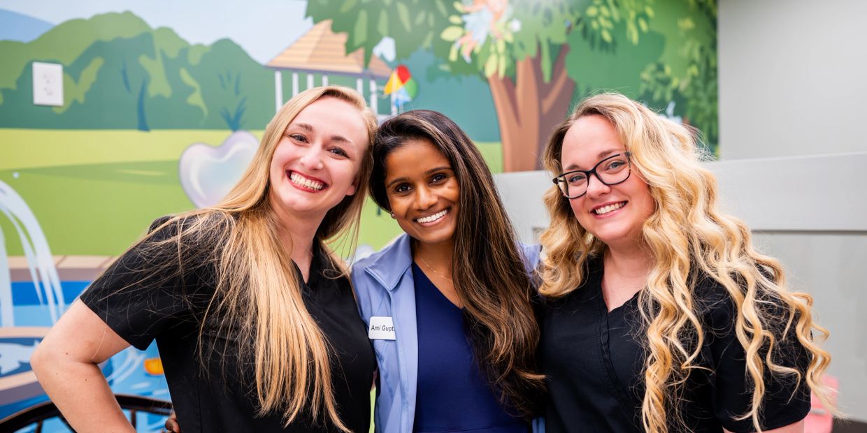 Three smiling women standing close together in front of a colorful mural.