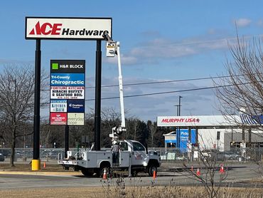 Worker in bucket truck repairing ACE Hardware sign on clear day.