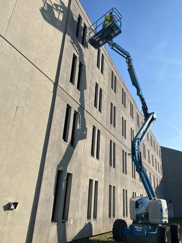 Worker on a blue boom lift inspecting a building wall with many windows.