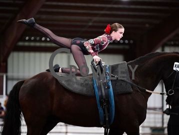 Young equestrian performing a gymnastic pose on a horse indoors.