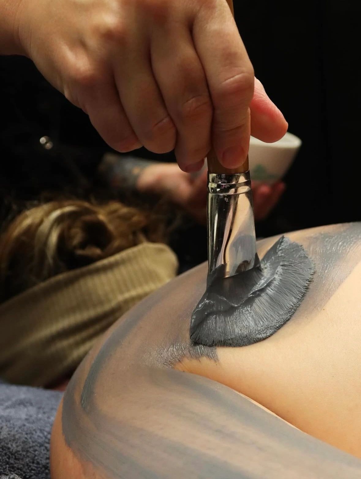 Close-up of a hand applying gray mud mask on skin with a brush.