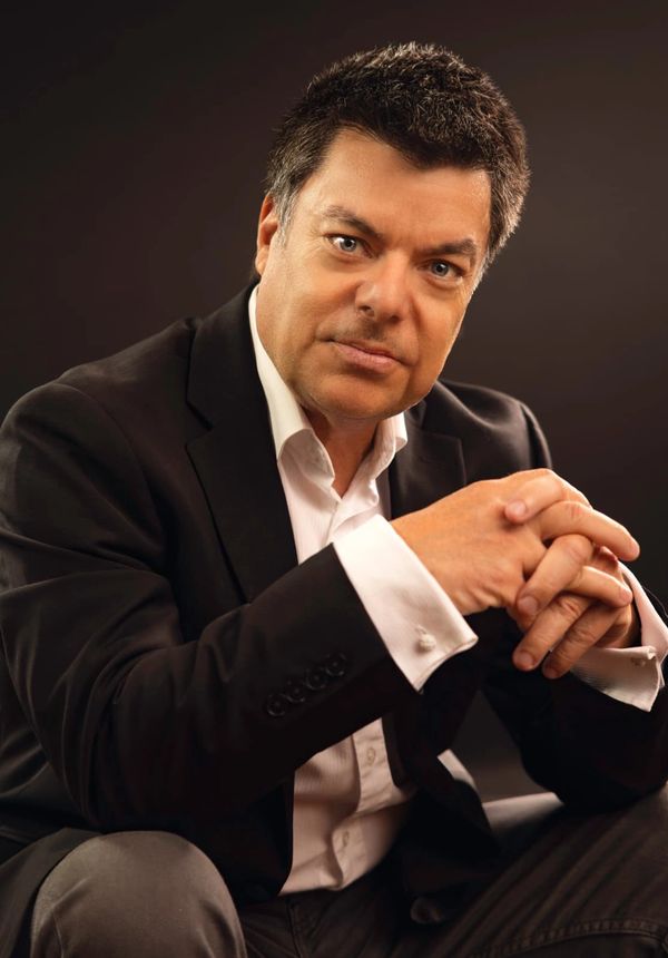 Confident man in a black suit posing thoughtfully against a dark background.