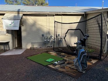 Outdoor golf practice setup with net, mat, and clubs beside a building.