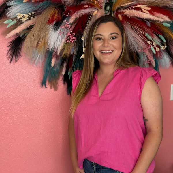 Smiling woman in a pink top against a pink wall with colorful pampas grass.