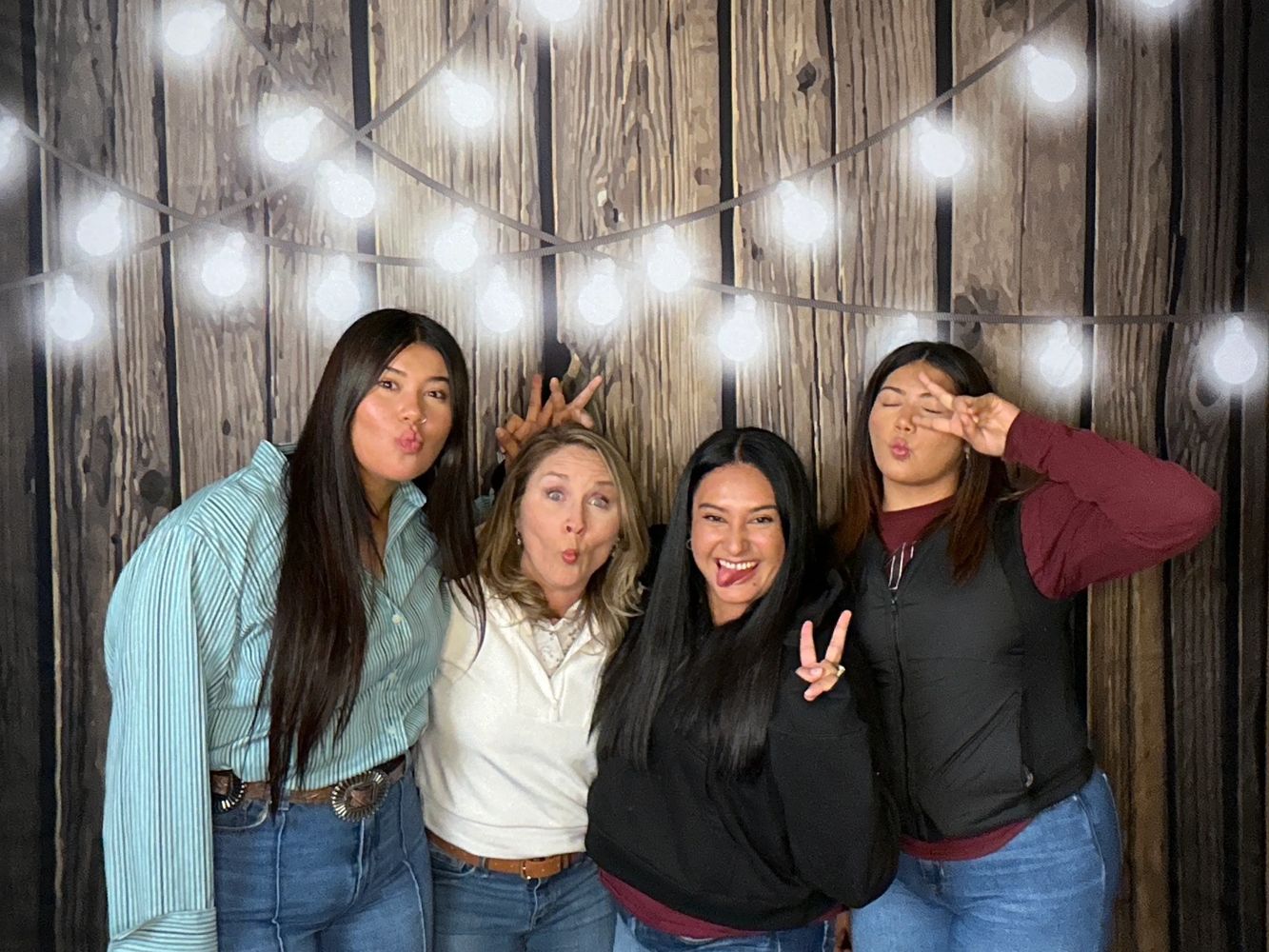 Four women posing playfully with peace signs and funny faces against a wooden backdrop with string lights.