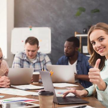A young woman gives a thumbs-up during a team meeting with laptops and documents.