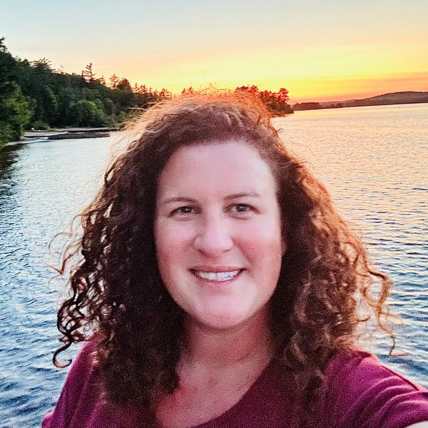 Woman with curly hair smiling by a lake at sunset.