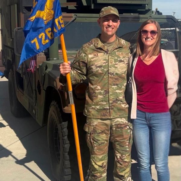 A soldier in uniform holding a flag stands next to a smiling woman in casual clothes.