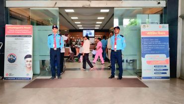 Two security guards stand at the entrance of a busy hospital lobby with people inside.