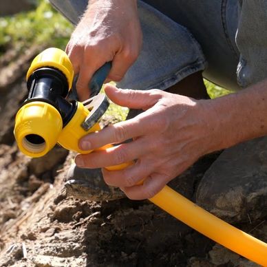 Person assembling a yellow plastic pipe fitting outdoors on dirt.
