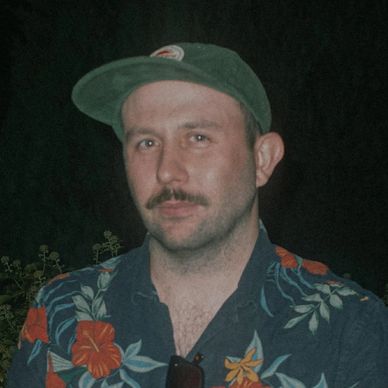 Man with a mustache wearing a floral shirt and green cap against a dark background.