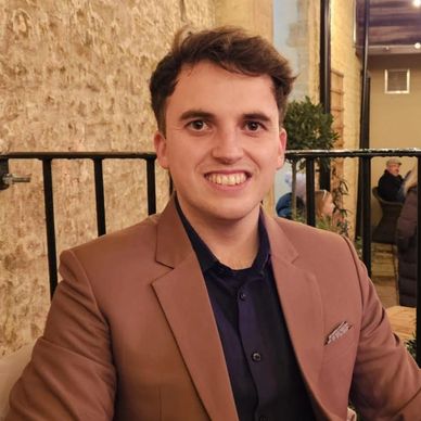 A young man smiling, dressed in a brown blazer and dark shirt, seated at an outdoor setting.