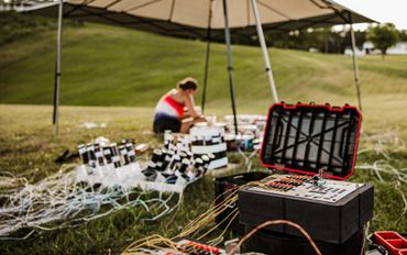 Person preparing fireworks setup with control panel outdoors under a canopy.