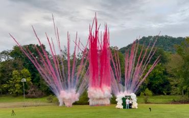 Colorful fireworks explode behind a couple standing in a balloon arch on a grassy field.