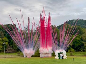 Pink fireworks explode behind a couple standing under a balloon arch outdoors.