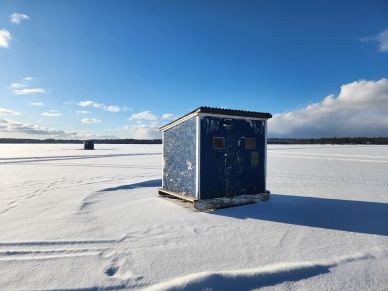 Blue ice fishing shack on a frozen snow-covered lake under clear sky.