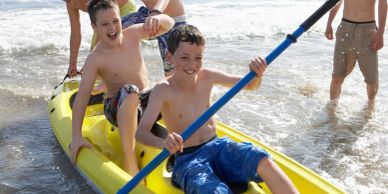 Two boys smiling and sitting in a yellow kayak on the shore.