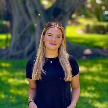 Young woman with long blonde hair stands outdoors in front of a large tree.