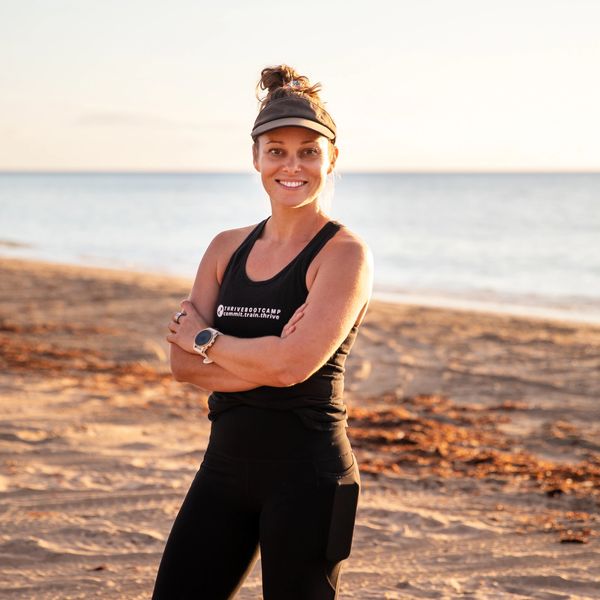 Smiling woman in workout attire stands confidently on a beach at sunset.