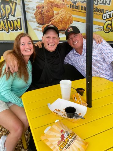 Three people happily posing at a yellow table with Crispy Cajun fried chicken food wrappers.